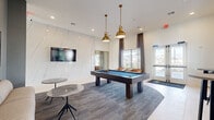 This expansive single-floor community clubhouse offers a well-rounded collection of amenity spaces designed for both relaxation and productivity. The building's exterior showcases a modern farmhouse aesthetic, combining white shiplap siding with stone-clad column bases, wood-look tile flooring on covered patios, and open views of the surrounding landscape and neighborhood.

Inside, two distinct living areas provide flexible gathering spaces. One features a full-size pool table beneath statement brass pendant lights, a dramatic white stone-accented accent wall, and a wall-mounted television, while the other offers lounge seating and additional TV viewing. The dedicated office space includes a built-in workstation with multiple computer terminals, brass industrial-style sconce lighting, and a textured accent wall, making it ideal for remote work or business needs.

The fitness center is a bright, well-equipped exercise room with striped performance flooring, ceiling fans, recessed linear lighting, and a variety of strength and cardio equipment. A dining room and connecting hallways complete the interior layout.

Outdoors, a generous covered porch runs the length of the building and is furnished with green cushioned seating and ceiling fans. A large resort-style swimming pool with in-water lounge chairs and surrounding deck areas serves as the centerpiece of the outdoor amenity experience.