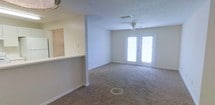 This single-floor space features an open-concept living area defined by soft, neutral-toned wall-to-wall carpeting and clean white walls that lend a bright, airy feel throughout. The living room is generously sized, providing ample space for comfortable furnishings and everyday living. A ceiling fan centered in the room adds both functionality and a practical touch to the space.

A standout feature is the set of French doors at the far end of the living area, which allow abundant natural light to flood the interior and offer direct access to an outdoor area. The doors serve as a focal point, enhancing the open, welcoming ambiance of the room.

To one side, a pass-through counter connects the living area to the adjacent kitchen, promoting an open flow between the two spaces. The counter is equipped with outlets, suggesting thoughtful utility placement. A glimpse of the kitchen beyond reveals light cabinetry and clean finishes. The textured ceiling adds a subtle visual element consistent throughout the space. Overall, the layout is practical and well-lit, suited for everyday comfortable living.