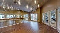This single-floor dance and fitness studio is thoughtfully designed to support a wide range of movement-based activities. The space features rich hardwood flooring that spans the entire room, providing a classic and durable surface ideal for dance, yoga, or group fitness classes. One wall is fully lined with floor-to-ceiling mirrors accented by a continuous wooden ballet barre, offering practitioners the ability to monitor form and technique during workouts or rehearsals. The warm, neutral-toned walls contribute to an inviting and focused atmosphere throughout the room. Elevated ceilings give the studio a spacious, open feel, allowing for unrestricted movement in all directions. Recessed and track lighting illuminates the space evenly from above, complemented by abundant natural light streaming in through a bank of large windows along one wall, which also offer views of a lush outdoor courtyard. A glass-paneled door provides a bright, welcoming entry point. Ceiling fans are mounted overhead to ensure comfortable air circulation during active use. The overall ambiance is clean, functional, and energizing, making this studio well-suited for dance instruction, personal training, or group exercise sessions.