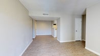 This single-floor space presents an open and airy layout that seamlessly connects the main living area to the kitchen beyond. Light hardwood-style flooring runs throughout, lending warmth and continuity to the space. The neutral, light gray walls are complemented by crisp white baseboards and trim, creating a clean, modern aesthetic.

The main living area is generously proportioned, offering ample room for a variety of furniture arrangements. A ceiling-mounted HVAC vent ensures year-round climate comfort. The open sightline extends toward a partially visible kitchen, which features under-cabinet lighting and a countertop pass-through or breakfast bar, adding both functionality and visual interest to the layout.

Two interior doors are visible — one leading to an adjacent room and another positioned along the far wall, likely providing access to additional spaces within the unit. Recessed lighting adds a subtle, ambient glow to the deeper portions of the space. Overall, the design emphasizes a bright, neutral palette and practical flow, making it a versatile and welcoming environment well-suited for everyday living.