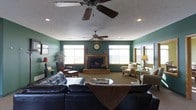 This single-story space centers around a well-appointed living room designed for comfort and relaxed gatherings. The room is finished with sage green walls that lend a warm, earthy tone throughout, complemented by neutral carpet flooring that adds to the inviting atmosphere.

A wood-framed fireplace serves as the focal point of the room, positioned along the main wall and flanked by multiple windows that allow generous natural light to filter in. The windows feature light-colored trim, providing a clean contrast against the green walls while offering views to the exterior.

Overhead, two ceiling fans are installed across the textured ceiling, ensuring comfortable airflow throughout the space. Recessed lighting supplements the natural light, contributing to a well-lit environment.

The living area is furnished with a substantial dark leather sectional sofa as its centerpiece, paired with a round coffee table. Additional seating, including upholstered armchairs and accent chairs, is arranged to encourage conversation. Decorative touches such as a wall clock, table lamps, and indoor plants complete the space, creating a cohesive and functional living environment.