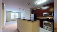 This single-floor residence offers a thoughtfully designed layout that balances functionality with a clean, modern aesthetic. The kitchen serves as a standout feature, showcasing rich dark mahogany cabinetry, sleek dark granite countertops, and a full suite of stainless steel appliances, including a refrigerator, electric range, and built-in microwave. Light tile flooring runs throughout the kitchen, and a peninsula with matching granite provides a natural divider between the kitchen and the adjoining living area. The open living space is carpeted in a neutral tone, creating a warm and comfortable atmosphere. A track lighting system illuminates the area, while a window with horizontal blinds allows natural light to filter in. A dedicated bedroom features plush carpeting, a ceiling fan, crown molding, and a window with an exterior view, contributing to an airy feel. The full bathroom is finished with tile flooring, a dark-cabineted vanity with a white countertop, a large mirror, and a tub-shower combination with tile surround. A separate utility area houses a stacked Whirlpool washer and dryer alongside a water heater, offering convenient in-unit laundry. Crown molding and consistent neutral wall tones unify the space throughout.