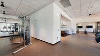 The fitness center at Local on Fourteenth is a thoughtfully designed, single-floor facility that offers a comprehensive workout environment for its users. The space is organized into several distinct zones, each tailored to a specific type of exercise.

Upon entry, a sleek reception area greets visitors with a dark cabinetry counter, floating shelves with decorative accents, white shiplap-style wall paneling, and speckled rubber flooring — a design detail that continues throughout much of the facility. Natural wood-framed glass doors provide a welcoming transition into the main workout areas.

The cardio zone is a standout feature, lined with floor-to-ceiling black-framed windows that offer expansive outdoor views of surrounding greenery. Rows of treadmills, ellipticals, stair climbers, and stationary bikes are positioned to take full advantage of this natural light and scenery. A ceiling fan overhead enhances comfort during workouts.

The strength training area provides ample space for cable machines and free weight equipment, complemented by recessed lighting and clean white walls. A dedicated studio room, enclosed by glass partitions and mirrored walls, features hardwood flooring and is equipped for group fitness, yoga, and functional training with stability balls, resistance tools, and yoga mats. The "Local on Fourteenth" branding is prominently displayed throughout the studio, reinforcing the facility's identity.