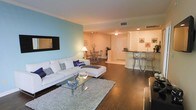 This single-floor residence showcases a thoughtfully designed open-concept layout that seamlessly connects the living, dining, and kitchen areas into one cohesive and functional space. Rich, dark hardwood flooring flows throughout, providing a warm and elegant foundation that ties the rooms together.

The living area is generously sized, anchored by a plush white sectional sofa and a sleek glass coffee table atop a textured area rug. A wall-mounted flat-screen TV is positioned against a striking teal accent wall, adding a bold pop of color that enlivens the space. Recessed and ambient lighting fixtures illuminate the room with a soft, inviting glow.

The dining area transitions naturally from the living room, featuring a compact dining table with stylish chairs set atop a patterned area rug, making it ideal for casual meals or entertaining.

The kitchen is defined by a raised breakfast bar with bar-stool seating, offering both a casual dining option and an open sightline into the main living space. Dark countertops contrast cleanly with the light cabinetry, creating a modern aesthetic. The open pass-through design ensures the kitchen feels integrated rather than isolated.

Overall, the space presents a modern, comfortable, and well-appointed interior with a cohesive design sensibility throughout.