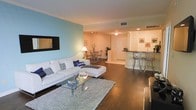 This single-floor residence showcases a thoughtfully designed open-concept layout that seamlessly connects the living, dining, and kitchen areas into one cohesive and functional space. Rich, dark hardwood flooring flows throughout, providing a warm and elegant foundation that ties the rooms together.

The living area is generously sized, anchored by a plush white sectional sofa and a sleek glass coffee table atop a textured area rug. A wall-mounted flat-screen TV is positioned against a striking teal accent wall, adding a bold pop of color that enlivens the space. Recessed and ambient lighting fixtures illuminate the room with a soft, inviting glow.

The dining area transitions naturally from the living room, featuring a compact dining table with stylish chairs set atop a patterned area rug, making it ideal for casual meals or entertaining.

The kitchen is defined by a raised breakfast bar with bar-stool seating, offering both a casual dining option and an open sightline into the main living space. Dark countertops contrast cleanly with the light cabinetry, creating a modern aesthetic. The open pass-through design ensures the kitchen feels integrated rather than isolated.

Overall, the space presents a modern, comfortable, and well-appointed interior with a cohesive design sensibility throughout.