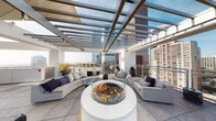 Located in the heart of downtown Los Angeles on S Olive Street, this striking rooftop amenity space seamlessly blends indoor comfort with outdoor living at an elevated vantage point above the city. The interior lounge is defined by a bold contrast of matte black ceilings and crisp white architectural columns, creating a modern, high-design atmosphere. Artistic pendant lighting with woven, sculptural shades adds warmth to the space, while large-format porcelain tile flooring runs throughout. Built-in banquette seating with plush cushions, woven rope dining chairs, and black glass-topped tables provide ample space for gathering and dining. A sleek wet bar with stainless steel fixtures, dark cabinetry, and reflective countertops serves as the central hub for entertaining. Floor-to-ceiling sliding glass walls open directly onto a spacious outdoor terrace, flooding the interior with natural light and framing unobstructed panoramic views of the Los Angeles skyline. The terrace itself is anchored by a circular fire pit surrounded by curved sectional seating, all sheltered beneath a modern pergola with glass-panel roofing. The result is an all-weather outdoor lounge ideal for gatherings, with the glittering downtown skyline as its backdrop.