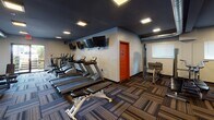 This fitness center is a thoughtfully designed, single-level workout facility offering a comprehensive range of exercise equipment and a modern aesthetic. The space is divided into distinct functional zones, providing dedicated areas for cardio training, strength work, and stretching or flexibility exercises.

The cardio zone features a row of treadmills and elliptical machines positioned along one wall, accompanied by wall-mounted flat-screen televisions for entertainment during workouts. Adjacent to this area, a strength training section includes a cable machine, adjustable weight bench, and a dumbbell rack stocked with a full range of free weights.

A large wall mirror spans one section of the facility, visually expanding the space and supporting proper form during exercise. A wooden ballet-style barre mounted in front of the mirror adds versatility for stretching and balance training.

The interior design combines dark, charcoal-painted ceilings with exposed industrial-style ductwork, giving the space a modern, urban character. Boldly striped accent walls in blue, orange, and teal add visual energy, while patterned multi-tonal carpet tiles cover the floor throughout. Recessed square lighting fixtures provide consistent illumination, and large windows along one end allow natural light and outdoor views into the space.