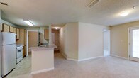 This single-floor residence offers an open-concept design that integrates the kitchen and living areas into one cohesive space. The kitchen features a peninsula counter with a light-toned laminate surface, an undermount sink, and wooden upper cabinetry in a warm medium-brown finish. A stainless steel refrigerator anchors the kitchen alongside additional counter space, providing functional meal preparation areas.

The living area is finished with wall-to-wall carpeting in a neutral beige tone, contributing to a warm and comfortable atmosphere. Smooth, neutral-painted walls run throughout the space, complemented by a textured ceiling with a centrally located HVAC ceiling vent, ensuring efficient climate control.

A hallway visible from the main living area leads toward what appears to be additional rooms or an entryway, indicated by a solid wood-style door at the far end. The layout flows naturally, making efficient use of the available footprint. Overall, the space presents a clean, understated aesthetic with practical residential finishes suited for everyday living.