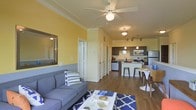 This single-floor residence showcases an open-concept layout that connects the living, dining, and kitchen areas into one cohesive and functional space. The living room is anchored by warm yellow accent walls contrasted with neutral white crown molding and trim, creating a cheerful and inviting atmosphere. A ceiling fan with a built-in light fixture serves as a central overhead feature, ensuring both comfort and illumination throughout the space. Hardwood-style flooring runs consistently across the entire open floor plan, adding warmth and visual continuity.

The kitchen is well-appointed with dark cabinetry, stainless steel appliances including a refrigerator, microwave, and range, and is complemented by pendant lighting that hangs above a breakfast bar with seating. Track lighting overhead provides additional task illumination. The dining area sits adjacent to the kitchen, offering a casual space for meals. A large mirror in the living area enhances the sense of openness. Overall, the space balances a lively color palette with practical design, making it both functional and visually engaging.