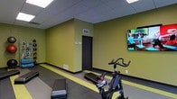 This single-level fitness space is designed to support a variety of cardio and exercise activities. The room features olive-green accent walls complemented by dark baseboards and a neutral drop-tile ceiling with recessed fluorescent lighting, creating a functional and energizing environment. A distinctive yellow floor stripe runs along the perimeter, adding a bold design element while visually defining the workout zone. The flooring consists of a combination of dark rubberized material and smooth surface sections, ideal for both equipment use and floor-based exercises. Key equipment includes a spin bike and step platforms, making the space suitable for cycling workouts and aerobic training. A wall-mounted flat-screen television displays workout content, supporting guided exercise sessions. Ventilation is provided through ceiling and wall-mounted HVAC vents, ensuring adequate airflow during intense workouts. A single door provides access to the room, keeping the layout open and unobstructed. The overall design is straightforward and practical, prioritizing functionality and ease of movement for users engaging in individual or small-group fitness activities.