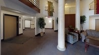 This expansive building lobby makes a strong first impression with its dramatic double-height ceilings and prominent structural columns that anchor the open atrium-style space. The main entrance features large glass doors that flood the foyer with natural light, leading into a generously sized common area finished with wall-to-wall carpeting and clean white walls accented by rich, dark wood trim details.

The lobby is thoughtfully arranged with a comfortable communal seating area, furnished with a plush sofa, upholstered armchairs, and a central coffee table — creating a welcoming space for residents to gather. Decorative indoor trees and potted greenery add warmth and life to the environment. Textile wall hangings draped from the upper level contribute an artistic, residential touch to the soaring interior.

An upper-level mezzanine with metal railings overlooks the main floor, reinforcing the building's verticality and open feel. A built-in mailbox station, set into a dark wood surround along one wall, provides a practical and centrally located amenity. A bulletin board nearby serves as a community information hub. Access doors throughout the lobby connect to additional corridors, offering clear circulation throughout the building. Overall, the space balances functionality with an inviting, community-oriented ambiance.
