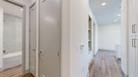 This well-appointed three-bedroom, two-bathroom residence in Austin, TX offers a thoughtfully designed single-floor layout with modern finishes throughout. The open-concept kitchen serves as the heart of the home, featuring a large quartz waterfall island, white shaker cabinetry extending to the ceiling, stainless steel appliances including a built-in microwave and oven, and a striking stainless steel range hood. Large windows flood the living and kitchen area with natural light and provide views of the surrounding Austin cityscape. Wood-look plank flooring flows throughout the main living areas and hallways, lending warmth and continuity to the space. The hallway features built-in shelving niches, adding functional storage and architectural interest. The primary bathroom is generously sized, offering dual vanities with white cabinetry and quartz countertops, wood-look flooring, and a glass-enclosed walk-in shower with tile surround. The secondary bathroom mirrors the same clean aesthetic with a glass-enclosed tub and shower combo. The bedrooms are carpeted for comfort, with ceiling fans and access to natural light. A private outdoor balcony with modern panel cladding, concrete flooring, and open views completes this contemporary Austin residence.