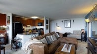 This single-floor residence offers an open-concept floor plan that brings together the living and kitchen areas in a cohesive, functional layout. The living room serves as the heart of the space, anchored by a large, plush sectional sofa in warm brown tones, complemented by decorative throw pillows and a sturdy wooden coffee table. Wall art and accent lighting add a personal, curated touch to the living area. The kitchen is well-appointed with rich medium-toned wood cabinetry, dark countertops, and black appliances including a refrigerator, microwave, and range. A breakfast bar with seating creates a casual dining option and reinforces the open connection between cooking and living spaces. The textured ceiling adds architectural character throughout, while flush-mount lighting ensures the space remains bright and welcoming. The neutral wall tones provide a versatile backdrop that enhances the warmth of the wood cabinetry and the earthy hues of the furnishings. Overall, the space delivers a comfortable and practical living environment with a cozy, lived-in ambiance suited for everyday life.