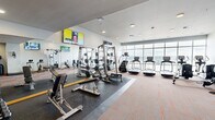 Located at 242 Broadway in Tacoma, Washington, this fitness center offers a thoughtfully designed single-floor workout facility with a comprehensive selection of exercise equipment. The space is organized into distinct functional zones, maximizing usability across its generous footprint.

The cardio zone is positioned along a floor-to-ceiling glass window wall that frames sweeping views of the surrounding skyline and waterfront, providing a visually stimulating backdrop for treadmills, stair climbers, ellipticals, and rowing machines. Natural light floods this area, creating an open and energizing atmosphere.

The strength training section features multiple cable and weight machines, a full rack of kettlebells and dumbbells, and adjustable benches set atop rubber flooring for safety and durability. Full-length mirrors line the walls, enhancing the sense of space while allowing users to monitor their form. Stability balls and a spin bike round out the functional fitness offerings.

Multiple wall-mounted flat-screen televisions are positioned throughout the room, keeping users entertained during workouts. Recessed ceiling lighting provides even, consistent illumination across the entire space. The flooring transitions between patterned carpet in the cardio and stretching areas and rubber matting in the strength zones. Warm wood-framed interior doors add a refined architectural touch to the overall clean, modern aesthetic.