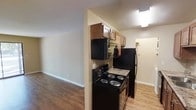 This single-floor space offers a practical and comfortable layout designed for everyday living. The kitchen is equipped with a full suite of black appliances, including an electric coil range, over-the-range microwave, and refrigerator. Medium-toned wood cabinetry provides ample storage overhead, while granite-look countertops add a touch of durability and style. A tiled backsplash accents the cooking area, complementing the overall warm, neutral color palette that runs throughout the space. The kitchen opens directly into the living area, creating a seamless flow between the two spaces. Wood-look laminate flooring extends across both areas, offering a cohesive and easy-to-maintain surface underfoot. The living area is bright and open, with clean white walls and white baseboards that enhance the sense of space. Recessed into the layout, a separate hallway leads to additional areas of the unit, suggesting a thoughtful floor plan that balances open communal spaces with more private zones. Overhead lighting provides consistent illumination throughout. The overall ambiance is clean, functional, and move-in ready, making it a well-suited space for comfortable day-to-day living.