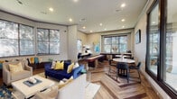 This well-appointed community lounge or clubroom is thoughtfully designed to accommodate both relaxation and social activity. The space is unified by a striking herringbone-patterned hardwood floor that adds visual depth and elegance throughout. Recessed lighting is embedded throughout the smooth ceiling, complemented by crown molding that frames each wall with a refined architectural touch.

The main lounge area features plush seating, including a bold navy blue velvet sofa accented with gold and patterned throw pillows, positioned around a central coffee table on a textured area rug. A standout corner nook offers built-in bench seating beneath a wrap of large windows fitted with warm wood blinds, lined with an abundance of decorative pillows — creating an inviting space for casual gathering or quiet reading. Adjacent decorative shelving displays curated accents alongside a textured wall panel.

A full-length shuffleboard table anchors the recreational zone, while nearby marble-top round tables paired with sleek black chairs provide a more intimate dining or conversation setting. Vibrant, original artwork adorns the walls, adding color and energy to the overall ambiance.

A wet bar area features dark wood cabinetry, a stainless steel sink, dishwasher, and light countertops beneath a window, offering functional amenity support. Large windows throughout allow ample natural light to fill the space, creating a bright and welcoming atmosphere.