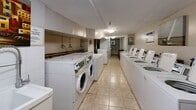 Located at 1276 Islington Ave in Toronto's Etobicoke neighbourhood, this shared laundry facility offers a practical and well-equipped space for residents. The room is laid out in a long, narrow corridor-style configuration, with rows of top-loading and front-loading washing machines lining both sides, complemented by stacked and standalone dryers. The machines are equipped with coin-operated or card-based payment terminals, making for a straightforward and convenient laundry experience. Underfoot, light-toned ceramic tile flooring runs the length of the room, providing a clean and easy-to-maintain surface. Overhead, flush-mounted ceiling fixtures provide consistent ambient lighting throughout the space. A wooden utility table is positioned along one wall, offering a dedicated surface for folding and sorting laundry. The room also includes a utility sink, posted informational signage, and a fire extinguisher, reflecting attention to safety and practicality. A secure entry door with a key access system ensures the facility remains accessible only to authorized users. The overall environment is functional, bright, and efficiently organized to accommodate the needs of a multi-unit residential building.