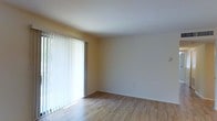 This single-floor space offers a clean and functional layout with a straightforward design. The main living area is highlighted by warm-toned hardwood flooring that runs throughout, lending a natural and inviting feel underfoot. A large sliding glass door serves as the room's focal point, fitted with full-length vertical blinds that allow for flexible light control while providing access to the outdoors. The generous size of the door draws in ample natural light, brightening the neutral, cream-colored walls and contributing to an open, airy ambiance. The walls are smooth and simply finished, providing a blank canvas well-suited for a variety of interior design styles. The ceiling is clean and white, further enhancing the sense of brightness and openness within the room. The overall aesthetic is minimal and practical, with an uncluttered layout that emphasizes space and ease of movement. The combination of natural light, hardwood flooring, and neutral tones creates a calm, versatile living environment ready to be personalized.