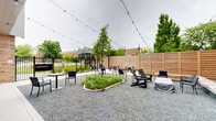 Located at 411 W Eighth St in Bloomington, Indiana, this thoughtfully designed outdoor courtyard offers a versatile communal space set between modern multi-story buildings featuring a mix of brick and paneled facades. The ground-level space is finished with gravel surfacing and concrete paver sections, providing distinct zones for relaxation and socializing. A central raised planter bed with ornamental grasses and young trees anchors the space, adding greenery and a natural focal point to the outdoor environment. String lights are strung overhead across the entire area, creating an inviting ambiance suitable for daytime and evening use. Seating options are varied and plentiful, including bistro-style round tables with chairs, Adirondack-style chairs, woven lounge chairs, and wooden picnic tables. A pergola structure on the perimeter adds architectural definition to one seating zone, while a covered fire pit provides an additional gathering feature. The courtyard is enclosed by a tall horizontal-slat cedar privacy fence on the street-facing sides, complemented by wrought iron fencing that maintains sightlines to the surrounding neighborhood and nearby greenery. Large glass windows and doors from the adjacent interior common area open visually onto the courtyard, creating a seamless indoor-outdoor connection. The space offers clear views of the Bloomington streetscape and lush tree canopy beyond the fencing.