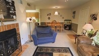This single-floor residence offers an open-concept layout that seamlessly connects the living, dining, and kitchen areas into one cohesive, functional space. The living area features plush carpeting and is anchored by a large, comfortable blue armchair complemented by a patterned area rug with fleur-de-lis motifs, adding a decorative focal point to the room. A floor lamp provides warm ambient lighting, enhancing the inviting atmosphere.

The dining area sits adjacent to the living space and accommodates a wooden table with matching chairs, illuminated by a classic pendant light fixture overhead. A two-tone wall treatment—neutral upper tones paired with a darker wainscoting-style lower section—runs throughout the open area, adding visual depth and architectural interest.

The kitchen is accessible via a pass-through counter, featuring warm wood cabinetry and tile flooring that transitions smoothly from the carpeted living and dining spaces. Decorative wall accents, including framed artwork and a fleur-de-lis motif, carry a consistent design theme throughout. An aquarium adds a lively, unique element to the space, contributing to the overall welcoming and personalized ambiance.