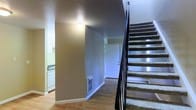 This two-story residence offers a practical and well-organized layout designed for comfortable everyday living. The main level features smooth hardwood-style flooring that flows through the entryway and into the adjacent spaces, creating a cohesive and easy-to-maintain foundation throughout. A carpeted staircase with a sleek black metal railing connects the ground floor to the upper level, serving as a central architectural element that adds visual contrast to the neutral interior palette.

The entryway provides a welcoming transition into the home, with a front door offering direct access to the main living area. A wall-mounted vent on the lower level ensures climate control convenience throughout the space. The walls are finished in a warm neutral tone that complements the overall interior scheme and creates a clean, open feel.

Adjacent to the entry, a glimpse into the kitchen reveals white cabinetry and standard appliances, suggesting a functional cooking and storage space. The layout positions the kitchen within easy reach of the main living and entry areas, supporting an efficient flow between rooms.

The upper floor, accessible via the interior staircase, expands the overall footprint of the home and provides additional living or sleeping quarters. Together, the two levels offer a well-structured and versatile living environment suitable for a range of occupants and lifestyles.