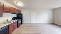 Located at 5609 Baker Ct in Virginia Beach, Virginia, this one-bedroom, one-bathroom unit offers a practical and well-organized single-floor layout. The open-concept kitchen and living area greets you upon entry through a classic six-panel door. The kitchen is equipped with rich cherry-toned cabinetry, light stone-look countertops, and a full suite of black appliances including a dishwasher, electric range, and refrigerator. Light wood-style flooring runs throughout the kitchen and living area, lending a clean, modern feel to the space. A hallway connects the main living area to the private quarters. The bedroom is a comfortable size, finished with soft wall-to-wall carpeting and neutral gray walls. A window fitted with blinds allows natural light to filter in, and a wall-mounted mini-split unit provides individual climate control. The bedroom also features a dedicated closet for additional storage. The bathroom includes a full-size soaking tub with white subway tile surround, complemented by light blue accent tile detailing along the lower wall, giving it a clean, classic aesthetic. Throughout the unit, consistent neutral tones and straightforward finishes create a cohesive, functional living environment.