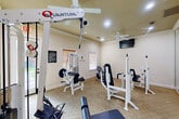 Located in Huffman, TX, this dedicated fitness center offers a well-equipped exercise space designed to support a comprehensive workout routine. The room is finished with warm, neutral-toned walls, crown molding, and carpeted flooring, creating a clean and inviting atmosphere for physical activity.

The space is outfitted with several Quantum-brand strength training machines, including cable pulley systems, a lat pulldown station, a leg press, and a seated exercise machine, providing users with a variety of resistance training options. A stationary recumbent bike adds a cardio element to the facility. A full-length wall mirror on one side of the room enhances the sense of space and allows users to monitor their form during workouts.

Natural light enters through a large glass-panel exterior door and a window fitted with blinds, both offering views of the surrounding green outdoor area. Recessed lighting and a ceiling fan with built-in light fixture ensure the space remains well-lit and comfortable. Two wall-mounted flat-screen televisions provide entertainment during workouts. A posted fitness room rules sign reflects the managed, community-oriented nature of the facility. The combination of functional equipment, thoughtful finishes, and practical amenities makes this fitness center a convenient and effective exercise destination.