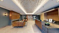 This well-appointed building lobby in Calgary, Alberta, makes a striking first impression through its refined architectural design and carefully curated finishes. The centerpiece of the space is an eye-catching geometric coffered ceiling adorned with a three-dimensional faceted panel that is elegantly backlit, casting a warm, ambient glow across the entire entrance area. Large-format neutral stone-look tiles extend throughout the floor, lending a clean and cohesive foundation to the space.

A dedicated reception desk, finished in rich wood tones with a dark stone countertop, anchors one side of the lobby and offers a professional welcome point for visitors. Adjacent to the desk, warm wood cabinetry and teal-painted millwork panels provide functional storage while adding visual depth and a refined color palette.

The lounge area invites guests to relax with a seating arrangement of cognac leather chairs and a sofa gathered around a central coffee table, all set upon a textured area rug. A sleek linear fireplace framed in stone and warm wood paneling serves as a focal feature in this seating zone, complemented by lush indoor plants and curated artwork.

Floor-to-ceiling glazed entry doors allow abundant natural light to flow inward, offering views of the surrounding streetscape. A connecting corridor, lined with bold abstract artwork and leading to the elevator bank, ensures a seamless and visually engaging circulation path through the lobby.