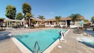 This outdoor recreational facility serves as a central amenity space within a residential community, designed to offer both leisure and accessibility. The centerpiece is a generously sized swimming pool with clear, blue-green water, complemented by a separate spa for relaxation. An ADA-compliant pool lift is installed at the pool's edge, ensuring the space is accessible to all residents and guests.

The surrounding pool deck is finished in smooth, durable concrete, providing ample space for lounge chairs and outdoor seating. A shaded patio area with a covered pergola structure offers a comfortable retreat from the sun, furnished with outdoor tables and chairs. Additional seating areas are scattered throughout the deck, ideal for socializing or unwinding.

The facility is enclosed by a secure wrought iron fence, enhancing safety while maintaining open sightlines to the lush landscaping beyond. Mature palm trees and shade trees frame the space, lending a warm, resort-like atmosphere. A stucco-clad amenity building with double French doors provides access to interior facilities, and an outdoor drinking fountain is conveniently positioned nearby. The overall design prioritizes comfort, accessibility, and a welcoming outdoor ambiance suited to warm-climate living.