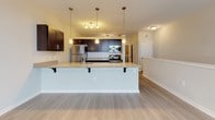 This single-floor residence is designed with a modern, open-concept layout that maximizes functionality and flow throughout. Light wood-tone flooring runs consistently through the main living areas, creating a cohesive and clean aesthetic.

The kitchen serves as a central feature of the space, showcasing rich dark cabinetry with sleek hardware, light granite countertops, and stainless steel appliances including a refrigerator, range, and microwave. A large peninsula with bar seating and pendant lighting above provides both additional prep space and casual dining options, while a track light fixture illuminates the cooking area effectively.

The living area is generously proportioned, offering ample room for furniture arrangements. A ceiling fan with a light fixture is centrally mounted, and large windows along one wall allow natural light to filter in while offering views to the exterior.

The bathroom features wood-look flooring consistent with the rest of the unit, a full tub/shower combination with built-in shelving, and neutral-toned textured walls. A dark vanity with a granite countertop completes the space.

A dedicated laundry room accommodates a full-size top-load washer and dryer side by side, with a wire shelving rack mounted above for additional storage convenience.