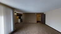This single-floor residential unit offers a straightforward, functional layout designed for comfortable everyday living. The main living area is finished with wall-to-wall carpeting in a neutral tone, creating a warm and cohesive foundation throughout the space. A textured popcorn ceiling adds a classic touch overhead, while a ceiling fan provides both lighting and air circulation.

The kitchen is positioned adjacent to the living area and is equipped with a white electric range, a refrigerator, and dark wood cabinetry overhead. A baseboard heater is visible along the wall, indicating an efficient heating setup suited for colder climates. The kitchen flows naturally into the main living space, making the overall floor plan feel open and connected.

A doorway visible from the living area leads to an additional room, which appears to include a closet with a rod, suggesting a bedroom or storage area. The walls are finished in a muted earthy tone, contributing to a neutral and understated interior palette throughout.

Overall, the space presents a practical and no-frills residential layout with all the essential areas accounted for, making it well-suited for those seeking a functional and easy-to-maintain living environment on a single level.