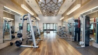 Located in Winter Park, Florida, this generously sized fitness center spans a single floor and is divided into two distinct areas designed to support a comprehensive workout experience. The main gym area immediately impresses with soaring ceilings adorned by a striking geometric lattice feature in warm gold tones, complemented by recessed lighting and modern pendant fixtures that create a bright, energizing atmosphere. Wood-plank flooring flows throughout, accented by rubber flooring zones in designated equipment areas. Floor-to-ceiling mirrors line the walls, enhancing the sense of openness and allowing members to monitor their form. The space is outfitted with an extensive array of strength training machines, free weights, cable systems, treadmills, ellipticals, and rowing machines, accommodating a wide range of fitness goals. Natural light pours in through large windows offering views of the surrounding outdoor landscape. A separate studio space serves as a dedicated cycling room, featuring a full wall of glass-panel garage doors that open to the outdoors, flooding the room with natural light and fresh air. This studio houses a row of cycling bikes alongside a wall-mounted display screen, making it well-suited for both individual and group sessions. Access between areas is provided through sleek glass-panel double doors, maintaining visual connectivity throughout the facility.