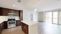 Located at 100 Citronica Ln in Lemon Grove, California, this single-floor unit offers a clean, modern aesthetic with thoughtfully designed living spaces. The open-concept kitchen is a standout feature, showcasing rich dark-wood cabinetry, stainless steel appliances including a range and microwave, and a central island with a crisp white countertop and gold-toned hardware, illuminated by elegant pendant lighting. The kitchen flows seamlessly into the living area, which is finished with light wood-look flooring that extends throughout the main living spaces, creating a cohesive and airy feel. A sliding glass door opens to a private balcony, allowing natural light to fill the living area and providing outdoor views of the surrounding community. The bedrooms feature plush carpeting and sliding closets with built-in rods for ample storage. The bathroom is well-appointed with a tiled soaking tub and shower combination, chrome fixtures, a dark-wood vanity with a white countertop, and a large mirror. Crisp white walls and clean lines throughout the unit contribute to a bright, contemporary ambiance that is both functional and inviting.