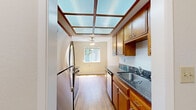 Located at 1137 Walpert St in Hayward, California, this single-floor, one-bedroom, one-bathroom residence offers a well-organized layout across a comfortable footprint. The galley-style kitchen is a standout feature, highlighted by a distinctive skylight panel ceiling that floods the space with diffused natural light. Warm wood cabinetry lines both sides of the kitchen, complemented by dark granite countertops, a stainless steel refrigerator, electric range, dishwasher, and a ceiling fan at the far end. Light hardwood-style flooring runs throughout the kitchen and hallway, adding a clean, cohesive feel. A short hallway connects the kitchen to the main living areas, with wood-panel interior doors providing access to the bathroom and additional storage. The living area is bright and open, featuring plush carpeting, a ceiling fan, and a large sliding glass door that opens to a private balcony with outdoor views of surrounding greenery. The generously sized bedroom offers carpet flooring, large windows with natural light and garden views, a built-in closet with shelving, and baseboard heating. The overall ambiance is warm and functional, with a neutral color palette throughout that lends itself to a variety of interior styles.