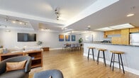 Located at 606 Third Avenue in the heart of downtown San Diego, this thoughtfully designed single-floor communal space offers a seamless open-plan layout integrating a living room, dining area, and kitchen. Light hardwood flooring runs throughout, creating a warm and cohesive aesthetic across the entire space.

The living room is generously sized, featuring large windows dressed with sheer white curtains that invite abundant natural light and offer views of the surrounding outdoor area. Comfortable seating arrangements, including a large sectional sofa and accent chairs, make this an inviting area for gathering and relaxation. A wall-mounted flat-screen TV and built-in shelving add functional appeal.

The dining area is anchored by a solid wood communal table surrounded by upholstered chairs, with a striking triptych of bold, colorful artwork adorning the wall above. Track lighting overhead highlights the space with a modern touch.

The kitchen is both stylish and practical, showcasing warm wood cabinetry, stainless steel appliances, and a large granite-topped peninsula with bar seating on multiple sides — ideal for casual dining or socializing. A luminous ceiling panel above the kitchen adds bright, even illumination. The neutral gray walls throughout provide a clean, contemporary backdrop that ties all areas of this versatile space together.