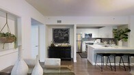 This single-floor space showcases a thoughtfully designed open-concept layout that seamlessly connects the living area and kitchen. The kitchen is well-appointed with stainless steel appliances, including a French door refrigerator, microwave, and range. Crisp white cabinetry paired with gray subway tile backsplash creates a clean, modern aesthetic, while a white-topped kitchen island with bar seating adds both functionality and a casual dining option.

Wood-style flooring runs throughout, providing warmth and continuity across the space. The living area features a neutral color palette with white walls, complemented by a textured ceiling. A recessed nook serves as a focal point between the kitchen and living area, housing a bold chalkboard-style wall art piece alongside a dark wood credenza, adding a decorative and functional element to the space.

The overall ambiance is contemporary and cohesive, with a contrast of dark and light tones creating visual interest. A doorway visible from the main living area suggests additional rooms beyond, contributing to the sense of a well-organized, flowing floor plan throughout the unit.