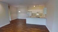 This single-floor space offers a clean, open-concept layout that seamlessly connects the living area and kitchen, creating a sense of spaciousness and easy flow between functional zones. The kitchen is appointed with crisp white cabinetry that extends to the upper walls, providing generous storage. A built-in microwave, electric range with a stainless sink, and a full-size refrigerator with an in-door water and ice dispenser complete the appliance package. The kitchen peninsula serves as a natural divider between the cooking and living areas, offering additional counter space and casual seating potential. Track lighting along the kitchen ceiling provides focused task illumination, while a pendant light fixture over the peninsula adds a warm, ambient glow. The living area is open and versatile, with neutral walls that offer a blank canvas for a variety of décor styles. Rich, dark hardwood flooring runs throughout the entire space, adding warmth and a sense of cohesion. Clean white baseboards provide a polished finish. The overall ambiance is bright, functional, and move-in ready, making it an ideal setting for both everyday living and casual entertaining.