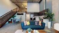 This two-floor loft showcases an industrial-modern aesthetic defined by exposed concrete ceilings, polished concrete floors, and raw structural elements throughout. The main level centers on an open-plan layout where the living, dining, and kitchen areas flow seamlessly together. The living room commands attention with dramatic double-height ceilings, an exposed concrete column, and floor-to-ceiling windows that flood the space with natural light and frame views of surrounding trees. A teal tufted sofa and mid-century modern furnishings reinforce the contemporary character of the space.

The kitchen features two-tone cabinetry—dark upper cabinets paired with white lower units—complemented by light countertops, a subway tile backsplash, and a full suite of stainless steel appliances. A dining area sits adjacent, completing the open-concept main level. Two bathrooms serve the space: one finished in crisp white with a shower-tub combination, and a second featuring bold navy walls, exposed ceiling pipes, and a floating white vanity. The bedroom continues the industrial theme with exposed concrete ceiling details and piping. A wood-railed staircase leads to the upper mezzanine level, adding functional versatility to this distinctly urban loft.