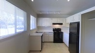 This single-floor space includes a well-appointed kitchen that serves as a central feature of the layout. The kitchen is designed with ample white cabinetry lining the upper and lower walls, providing generous storage throughout. Black appliances, including an electric range with a ventilation hood and a refrigerator, create a striking contrast against the white cabinetry and light-toned countertops. A sink is conveniently positioned near a window fitted with horizontal blinds, allowing natural light to fill the space while maintaining privacy. The light-colored tile flooring adds to the bright, clean aesthetic of the kitchen. Overhead, a track lighting system runs the length of the ceiling, offering adjustable, functional illumination across the workspace. The U-shaped kitchen layout maximizes counter space and promotes an efficient workflow. The neutral wall tones and understated finishes throughout contribute to a straightforward, practical ambiance. Overall, the space presents a clean, functional environment with a cohesive design centered on usability and simplicity.