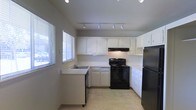 This single-floor space includes a well-appointed kitchen that serves as a central feature of the layout. The kitchen is designed with ample white cabinetry lining the upper and lower walls, providing generous storage throughout. Black appliances, including an electric range with a ventilation hood and a refrigerator, create a striking contrast against the white cabinetry and light-toned countertops. A sink is conveniently positioned near a window fitted with horizontal blinds, allowing natural light to fill the space while maintaining privacy. The light-colored tile flooring adds to the bright, clean aesthetic of the kitchen. Overhead, a track lighting system runs the length of the ceiling, offering adjustable, functional illumination across the workspace. The U-shaped kitchen layout maximizes counter space and promotes an efficient workflow. The neutral wall tones and understated finishes throughout contribute to a straightforward, practical ambiance. Overall, the space presents a clean, functional environment with a cohesive design centered on usability and simplicity.