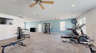 This spacious fitness center is thoughtfully designed to accommodate a wide range of workout routines within a single open floor plan. The interior is divided into distinct functional zones, allowing for both cardiovascular and strength training activities without feeling overcrowded. Walls are finished in a calming combination of soft blue and crisp white, creating an energizing yet comfortable atmosphere throughout.

The cardio area is well-stocked with treadmills, elliptical machines, and stationary cycling bikes, each positioned near large windows that allow natural light to fill the space. Wall-mounted televisions are strategically placed for viewing during workouts. Multiple ceiling fans provide consistent air circulation, supplemented by recessed lighting overhead.

The strength training section features a multi-functional cable machine, a comprehensive dumbbell rack with an adjustable weight bench, and additional resistance equipment. A full-length wall mirror in the free weights area enhances the sense of space and supports proper form during exercise. A water fountain station is conveniently located nearby.

Durable, textured carpet tile covers the floors throughout, offering both comfort and practicality. The open layout provides generous circulation space between equipment, and a storage unit offers organized space for accessories. Overall, the fitness center presents a clean, functional, and well-maintained environment suited for diverse fitness needs.