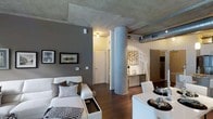 This single-floor urban loft showcases a compelling blend of industrial and contemporary design. The open-concept living area is defined by a raw exposed concrete ceiling and visible silver ductwork, lending an authentic industrial character throughout the space. A prominent cylindrical concrete column anchors the layout, serving as a striking architectural focal point that separates the living and dining areas while maintaining an open, airy feel.

Rich dark hardwood flooring runs throughout, providing a warm contrast to the cool concrete overhead. The living area features a modern white sectional sofa accented with layered throw pillows, set against a bold charcoal feature wall adorned with framed artwork. Recessed and track lighting illuminate the space, complementing the industrial aesthetic.

The dining area is furnished with a sleek white table and matching chairs, positioned near a decorative console that adds an artistic touch. Adjacent to the main living space, a hallway leads to additional rooms, with clean white doors and walls offering a crisp, transitional contrast to the open loft's edgier design language. A glimpse of cabinetry hints at a functional kitchen nearby. Overall, the space delivers an urban, loft-style ambiance with thoughtful modern furnishings and cohesive design details.