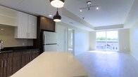 Located at Bishops, this single-floor residence offers a thoughtfully designed open-concept layout that seamlessly connects the kitchen and living areas. The contemporary kitchen is a standout feature, showcasing rich dark wood cabinetry that contrasts elegantly with crisp white upper cabinets and a stainless steel refrigerator. A light-toned countertop island serves as a central gathering point, ideal for meal preparation or casual dining. Overhead, a combination of stylish pendant lighting and adjustable track lighting ensures the space is both functional and atmospherically lit. The open living area extends naturally from the kitchen, bathed in abundant natural light streaming through expansive floor-to-ceiling windows. These large windows not only brighten the interior but also frame views of the surrounding outdoor environment, creating a seamless connection between inside and out. Light hardwood-style flooring runs throughout, adding warmth and continuity to the space. A recessed ceiling detail in the living area adds architectural interest and subtle definition between zones. A doorway visible from the main living space suggests additional rooms beyond, contributing to the overall functional flow of the layout. The overall ambiance is clean, modern, and bright.