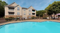 This outdoor pool area offers a well-designed communal retreat centered around a generously sized swimming pool with clear, blue water and tiled edges. The surrounding concrete sundeck provides ample space for relaxation and is furnished with multiple adjustable lounge chairs, ideal for sunbathing or unwinding outdoors. A covered patio area supported by stacked-stone columns provides a shaded gathering space complete with outdoor dining furniture, offering a comfortable retreat from direct sunlight. The pool area is enclosed by a wrought-iron fence, providing both security and an open, airy feel. Restroom facilities are conveniently accessible directly from the pool deck, housed within a structure featuring distinctive stacked-stone masonry accents and large multi-pane windows that offer views of the pool area. The building backdrop is a multi-story residential structure with neutral tan siding, stone accents, gabled rooflines, and private balconies that overlook the pool. Mature trees surrounding the perimeter add natural greenery and shade, enhancing the overall ambiance. The entire space benefits from abundant natural light and open sky views, creating an inviting outdoor environment suited for both active recreation and leisurely relaxation.