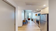 This single-floor unit is designed with a clean, contemporary aesthetic that blends functionality with modern style. The open-plan layout connects the kitchen, dining, and living areas in one continuous space, creating a streamlined flow ideal for daily living. Light hardwood flooring runs throughout, complementing the bright white walls and exposed concrete structural columns that add an industrial character to the interior.

The kitchen is well-appointed with light wood cabinetry extending to the ceiling for ample storage, stainless steel appliances including a full-size refrigerator, electric range, built-in microwave, and a single-basin sink. Recessed lighting illuminates the kitchen and living areas effectively. The living space accommodates a sofa and seating arrangement, with a dining table and chairs positioned nearby.

The bedrooms are thoughtfully positioned to maximize the building's elevated vantage point, each featuring floor-to-ceiling windows that frame sweeping panoramic views of the surrounding cityscape, waterway, and urban skyline. Exposed concrete columns appear throughout, lending architectural continuity. The bedroom with corner windows offers an especially commanding view. Built-in under-bed storage drawers provide practical space solutions. Natural light floods every room, enhancing the open and airy ambiance of this well-designed urban residence.