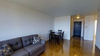 This single-floor space combines a living and dining area into one cohesive, open-concept room. The flooring throughout is a standout feature — warm-toned parquet wood arranged in a classic herringbone-style pattern, adding texture and character to the interior. The walls are finished in a clean, neutral white, contributing to a bright and airy atmosphere that makes the space feel open and welcoming.

The living area is furnished with a tufted grey sofa accented by a decorative geometric-patterned cushion, offering a comfortable seating arrangement. Adjacent to this, the dining area accommodates a dark wood table paired with matching chairs, providing a practical space for everyday meals. A partial wall subtly defines the transition between these two zones while maintaining an open feel.

A hallway visible from the main living area leads toward additional rooms, with a ceiling-mounted light fixture providing illumination along the corridor. The entry door is finished in a deep charcoal tone, creating a subtle contrast against the lighter interior. Overall, the space presents a clean, functional layout with warm flooring details and a simple, understated aesthetic.