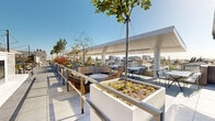 This rooftop amenity level offers a thoughtfully designed combination of indoor and outdoor spaces, blending contemporary architecture with lush greenery and sweeping views of the surrounding cityscape and distant mountains. The outdoor terraces are generously proportioned, featuring large-format porcelain tile flooring and an array of raised planters filled with seasonal plantings, succulents, and mature trees that provide natural shade and a sense of enclosure. Multiple seating configurations are distributed throughout—including intimate lounge chairs, wicker accent seating, and bistro-style table-and-chair sets—accommodating both casual gatherings and larger social occasions. String lighting overhead adds an ambient quality to the space after dark. A partially covered pergola structure with clean white columns extends over a portion of the terrace, offering shade while maintaining open-air connectivity. A secondary terrace with a built-in outdoor kitchen or grill station provides additional functionality. The interior clubroom is framed by floor-to-ceiling glass on multiple sides, flooding the space with natural light and framing unobstructed views of the skyline. Inside, neutral tones, sheer drapery, polished tile floors, and contemporary furnishings create a refined yet approachable atmosphere suited for gatherings or quiet retreat.