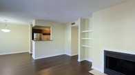 This single-floor space offers an open and functional layout that maximizes the sense of room. The living area features rich, dark hardwood flooring that contrasts cleanly with the light cream-toned walls and bright white baseboards, creating a classic and neutral aesthetic throughout. A textured popcorn ceiling adds a traditional finish overhead, while a ceiling vent ensures efficient climate control.

A standout feature of the living area is the built-in wall shelving unit, offering three open shelves ideal for display or storage. Adjacent to the shelving, a fireplace adds a focal point to the room, enhancing both functionality and ambiance.

The kitchen is accessible via a pass-through opening from the living area, promoting an open-concept feel while maintaining a defined cooking space. The kitchen is equipped with a stainless steel refrigerator, warm wood cabinetry, a mosaic tile backsplash, and a countertop that doubles as a breakfast bar. An overhead range hood is mounted above the cooking area.

Overall, the space presents a clean, move-in-ready environment with thoughtful built-in features and a cohesive, neutral design palette suitable for a variety of furnishing styles.