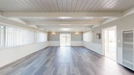 Located at 13475 Bancroft Avenue in San Leandro, CA, this single-floor space offers a thoughtfully designed, open-plan living area that balances modern finishes with classic architectural charm. The room is defined by its striking white-painted exposed beam ceiling with shiplap detailing, which adds both character and a sense of elevated height to the interior. Neutral gray-toned luxury vinyl plank flooring runs throughout, providing a clean, contemporary foundation that complements the soft greige wall tones. A classic white-mantled fireplace serves as a natural focal point on one wall, lending warmth and a timeless aesthetic to the space. White wainscoting lines the lower portion of the walls, adding a refined architectural detail that ties the interior together. Natural light is a defining feature, streaming in through multiple large windows fitted with vertical blinds, as well as a sliding glass door that opens to the outside. Wall-mounted sconce lighting adds a warm ambient glow. A wall-unit HVAC system ensures year-round climate comfort. The open layout offers exceptional flexibility, making the space well-suited for a variety of uses. Overall, the space presents a clean, bright, and welcoming atmosphere with a cohesive blend of traditional and contemporary design elements.