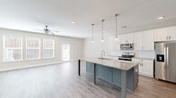 Located at 133 Dolcetto Dr in Winder, Georgia, this two-story home offers four bedrooms and three bathrooms thoughtfully distributed across two floors. The main level opens into a bright, open-concept kitchen and living area finished with light wood-look flooring and a neutral color palette. The kitchen is a standout, featuring a large center island with a striking teal base, granite-look countertops, a farmhouse-style sink, pendant lighting, crisp white cabinetry, and stainless steel appliances including a refrigerator, range, and microwave. The adjacent living room is bathed in natural light from multiple windows and a rear door, with a ceiling fan overhead. A pantry and hallway round out the main floor's functional layout. Carpeted stairs with a white handrail lead to the upper level, which houses all four bedrooms, including a generously sized primary suite with a walk-in closet fitted with wire shelving. The secondary bedrooms feature plush carpet, ceiling fans, and windows that invite natural light. A dedicated laundry room on the upper floor includes a side-by-side Whirlpool washer and dryer with additional shelving. Three bathrooms serve the home, each finished in clean, neutral tones. The home's design balances practicality with modern style throughout.
